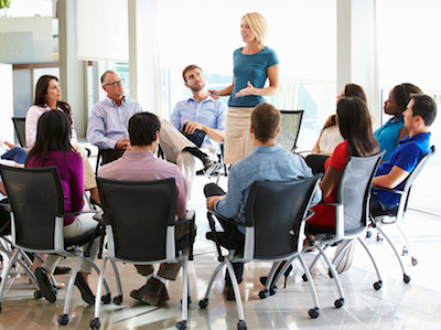 a young woman speaks to a group sitting in a circle in a warmly lit room